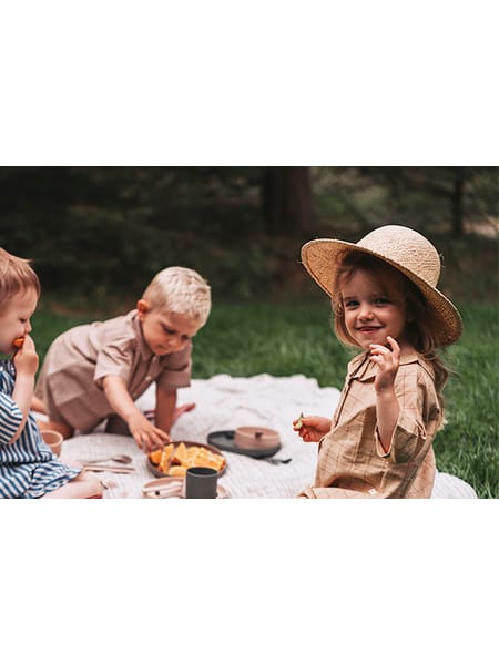 Børn og voksen på tæppe i haven til picnic, med legetøj og mad på en solrig dag.
