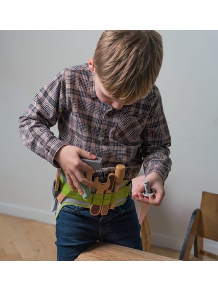 Et barn med værktøjsbælte står ved et træbord og ser ned på noget i sine hænder.