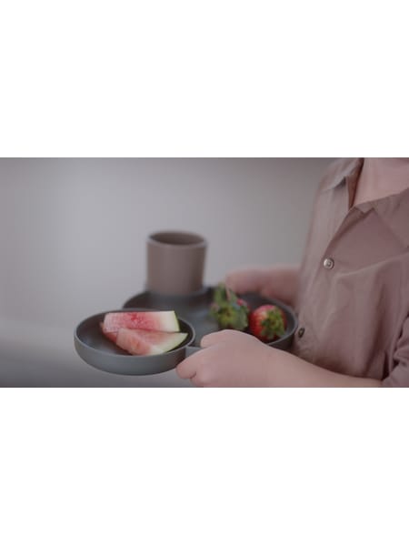 En person holder en kop og tallerken i lyserødt med små grøntsager ved siden af på en grå bakke.