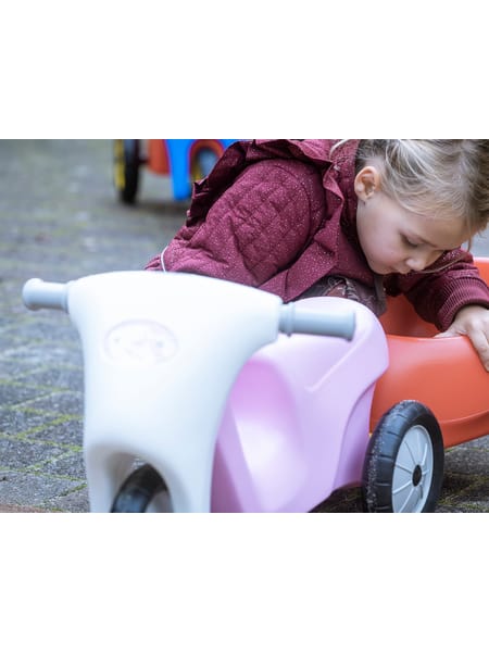 Barn på hvidt plastiksæde med hjul på grå asfalt, iklædt bordeaux jakke og brune støvler