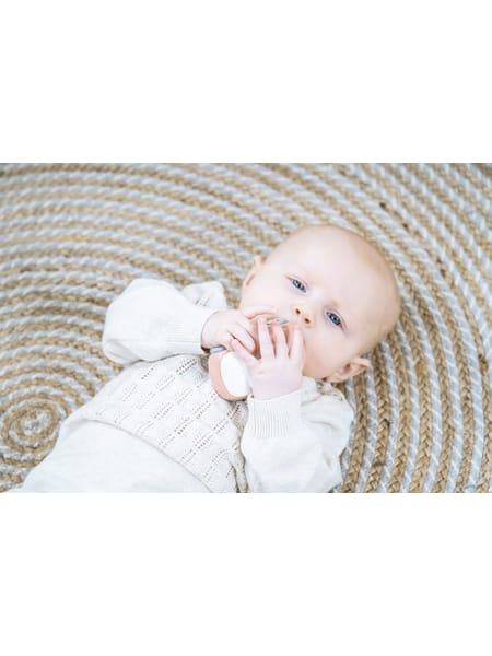 Baby i hvid kjole ligger på rundt, beige tæppe mens barnet rører ved sin mund