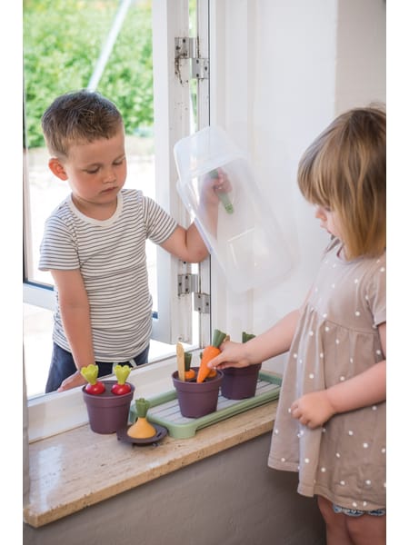 To børn ved træbord leger med havesæt og vandkande i solrigt vejr med stakit i baggrunden