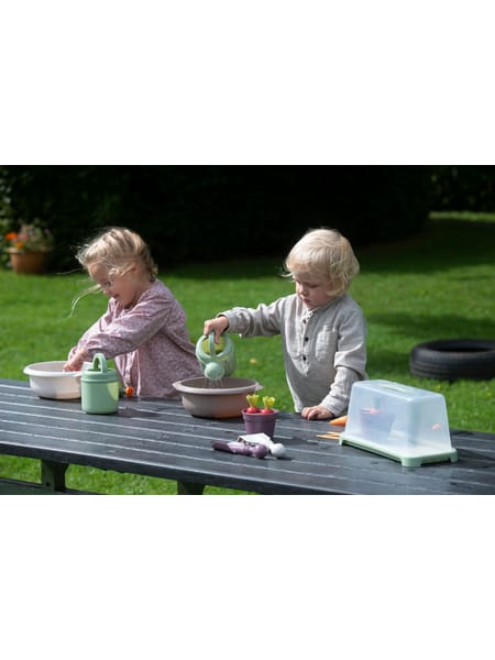 To børn leger med planteskolesæt ved havebord udendørs med grøn græsplæne i baggrunden