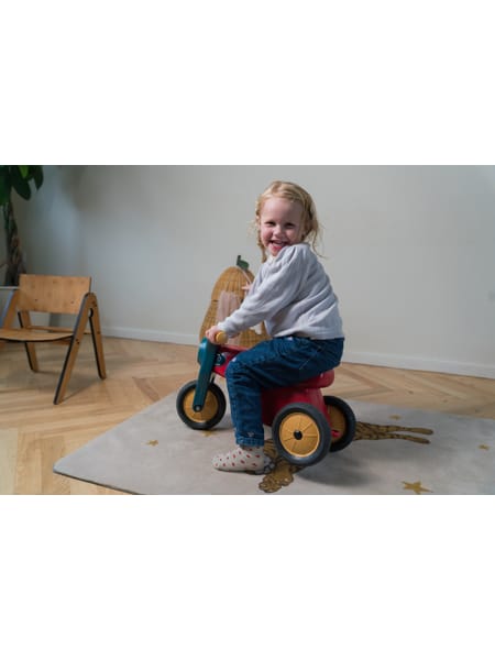 Barn på trehjulet motorcykel i blå og gul på trægulv ved stol