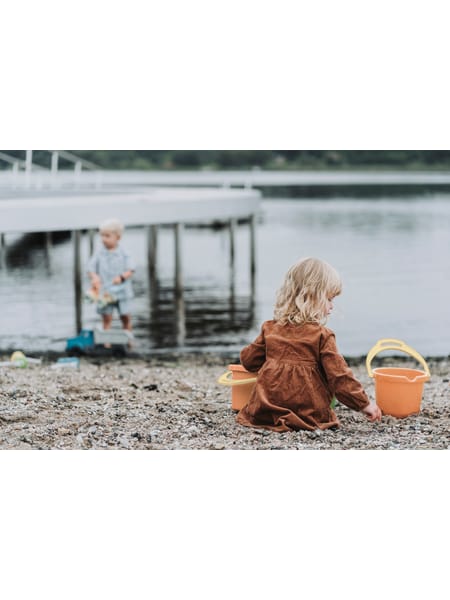 Barn i brun frakke med orange spand sidder ved vandet på stenet strand med flydebro i baggrunden