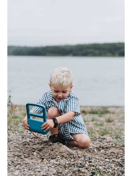 Lille barn med blåt sandlegetøj ved vandkant på en sommerdag.