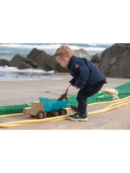Barn leger med blå dumper på stranden ved siden af grønt legetøj og klipper.