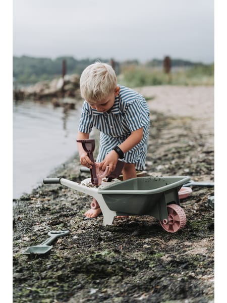 Barn med grøn trillebør leger ved stranden i gråvejr