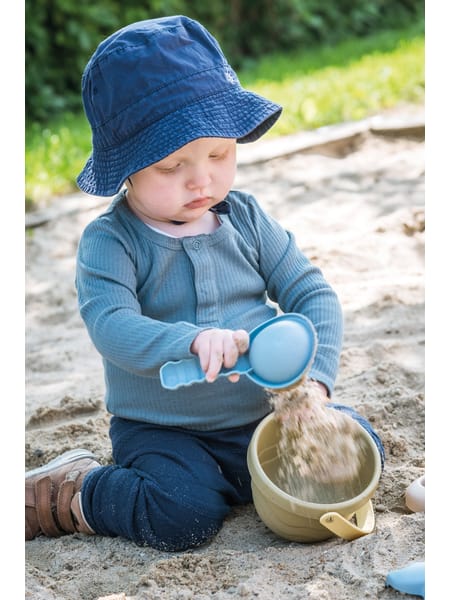 Lille barn i blå tøj og hat sidder udendørs og leger med et spand i lysebrun farve.