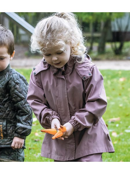 Barn med orange haveredskab leger udenfor i en brun jakke på en grøn græsplæne.