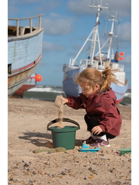 Ved stranden leger et barn med spand og legetøj mens fiskerbåde ses i baggrunden