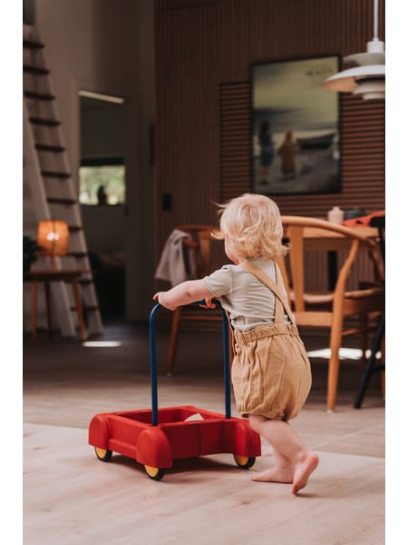Lille barn trækker rød trækvogn på hjul henover et trægulv i et hjemligt miljø