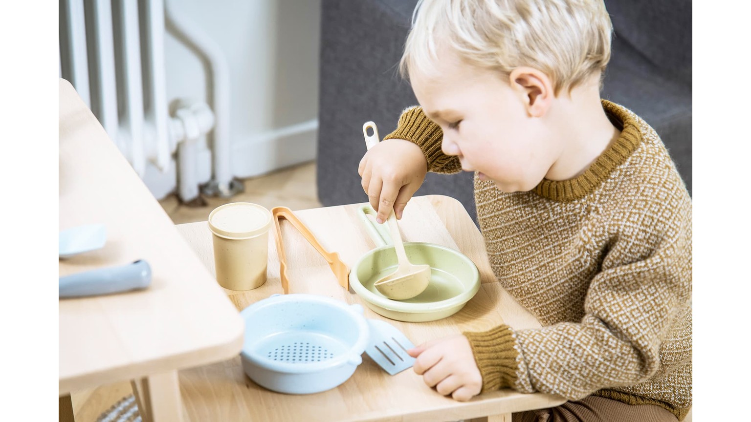 Barn i brun sweater spiser af grøn skål ved bord med blå skål ved siden af.