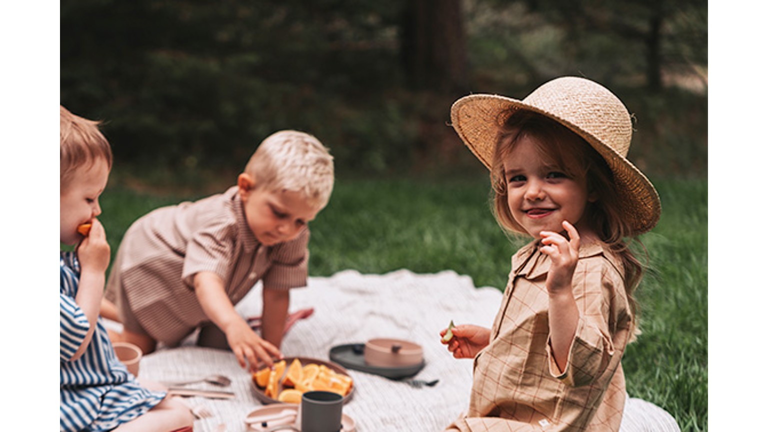 Børn og voksen på tæppe i haven til picnic, med legetøj og mad på en solrig dag.