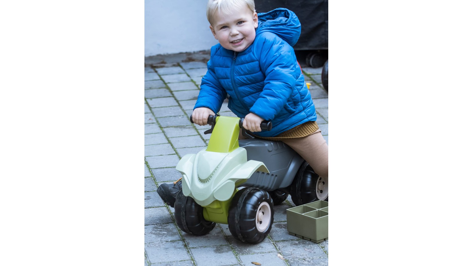 Et barn i blå jakke kører på et lille hvidt og grønt legetøjs-ATV på flisebelagt underlag