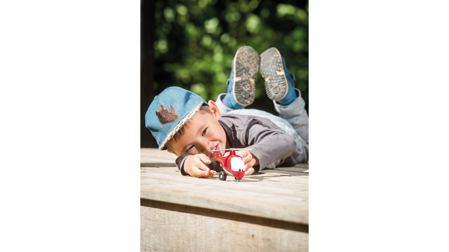 Lille person med blå kasket ligger på siden og leger med en rød bil på træoverflade.