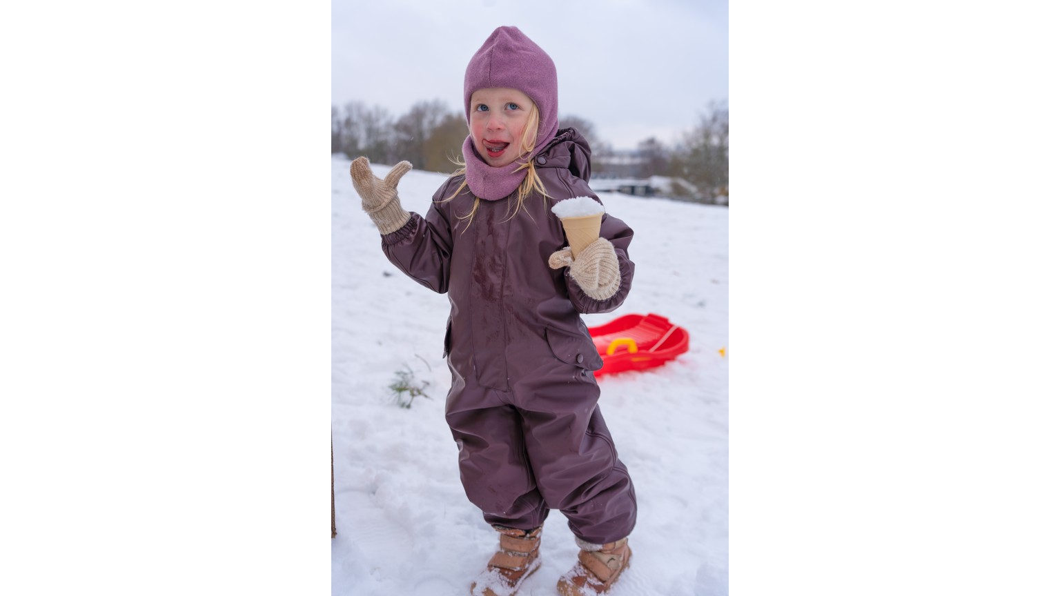 Barn i brun vinterdress leger i sneen med en rød plastslæde