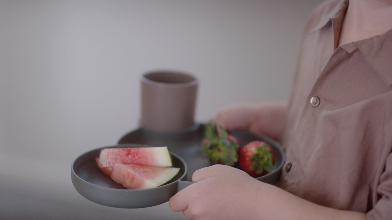 En person holder en kop og tallerken i lyserødt med små grøntsager ved siden af på en grå bakke.