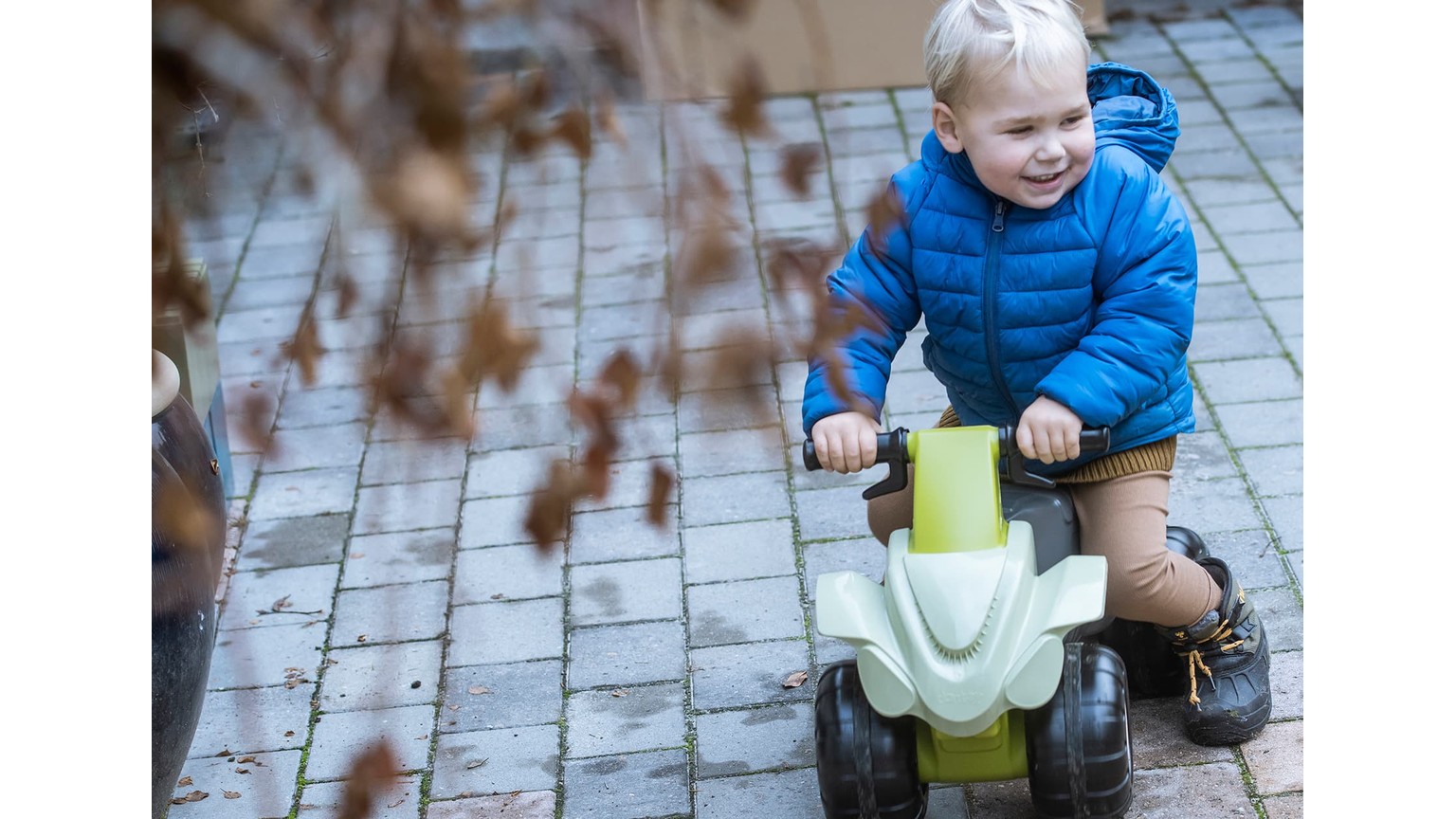 Barn på blå plastik-ATV på flisebelagt område med efterårsgrene i forgrunden