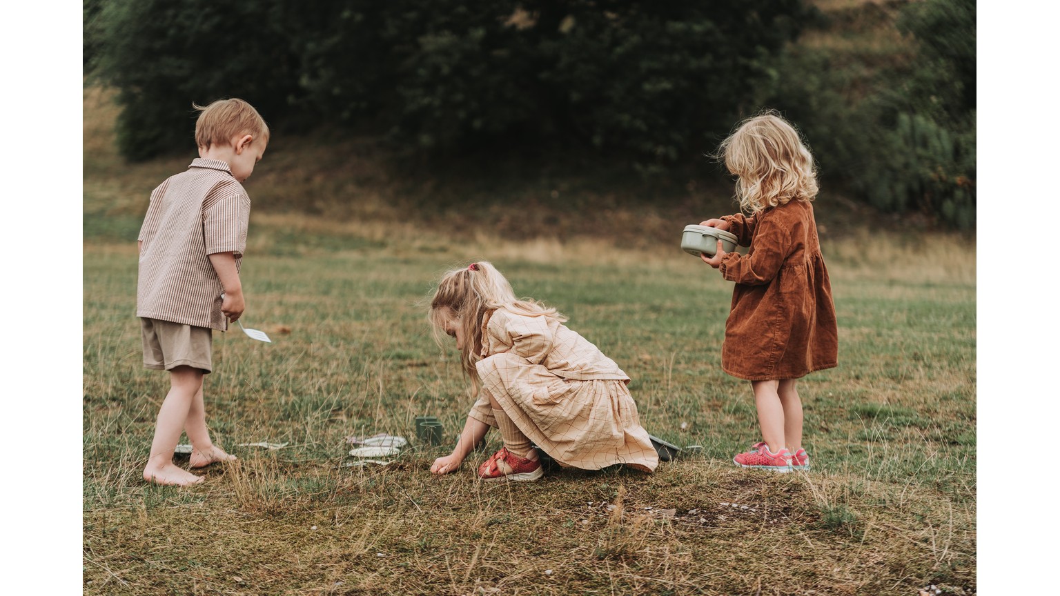 Tre børn leger i det høje græs på en mark i naturlige efterårsfarver.