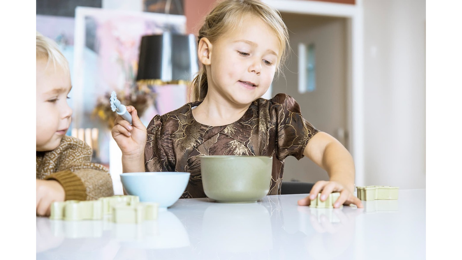 To børn ved et bord med skåle foran sig under madlavningsaktivitet i hjemligt miljø.