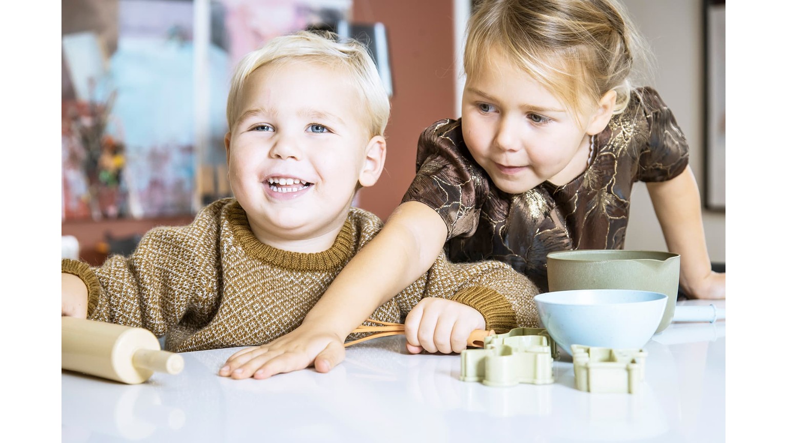 To børn sidder ved et bord med bageredskaber og skåle foran sig i en hyggelig indendørs setting.