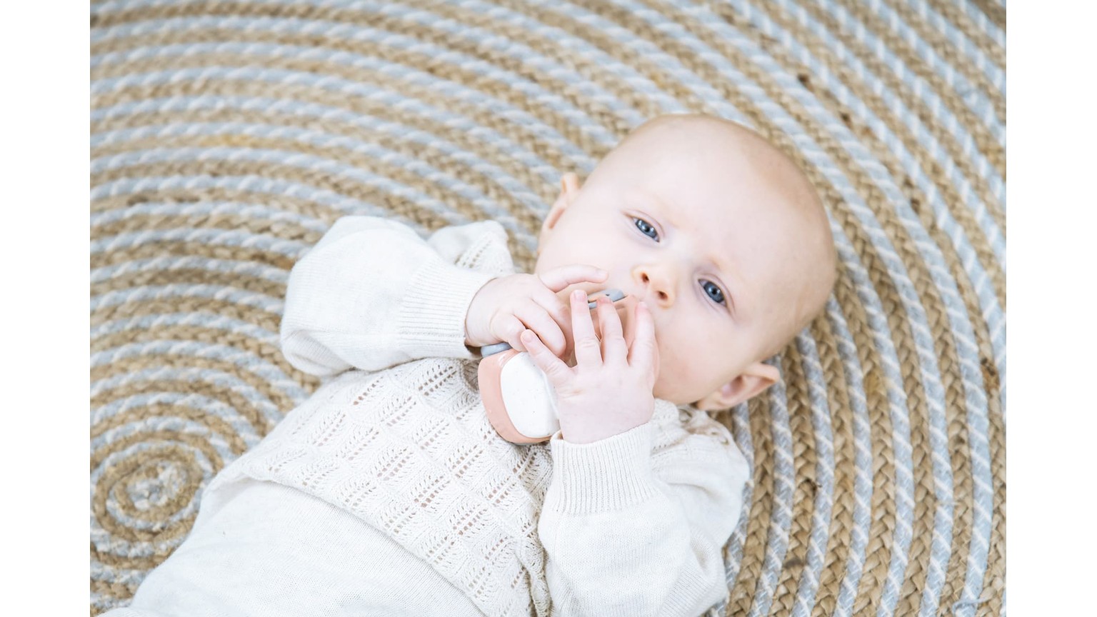 Baby i hvid kjole ligger på rundt, beige tæppe mens barnet rører ved sin mund