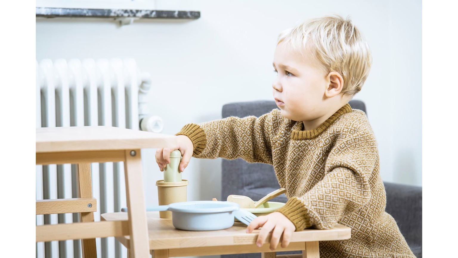 Barn ved bord med skål og ske spiser morgenmad ved siden af en hvid radiator.
