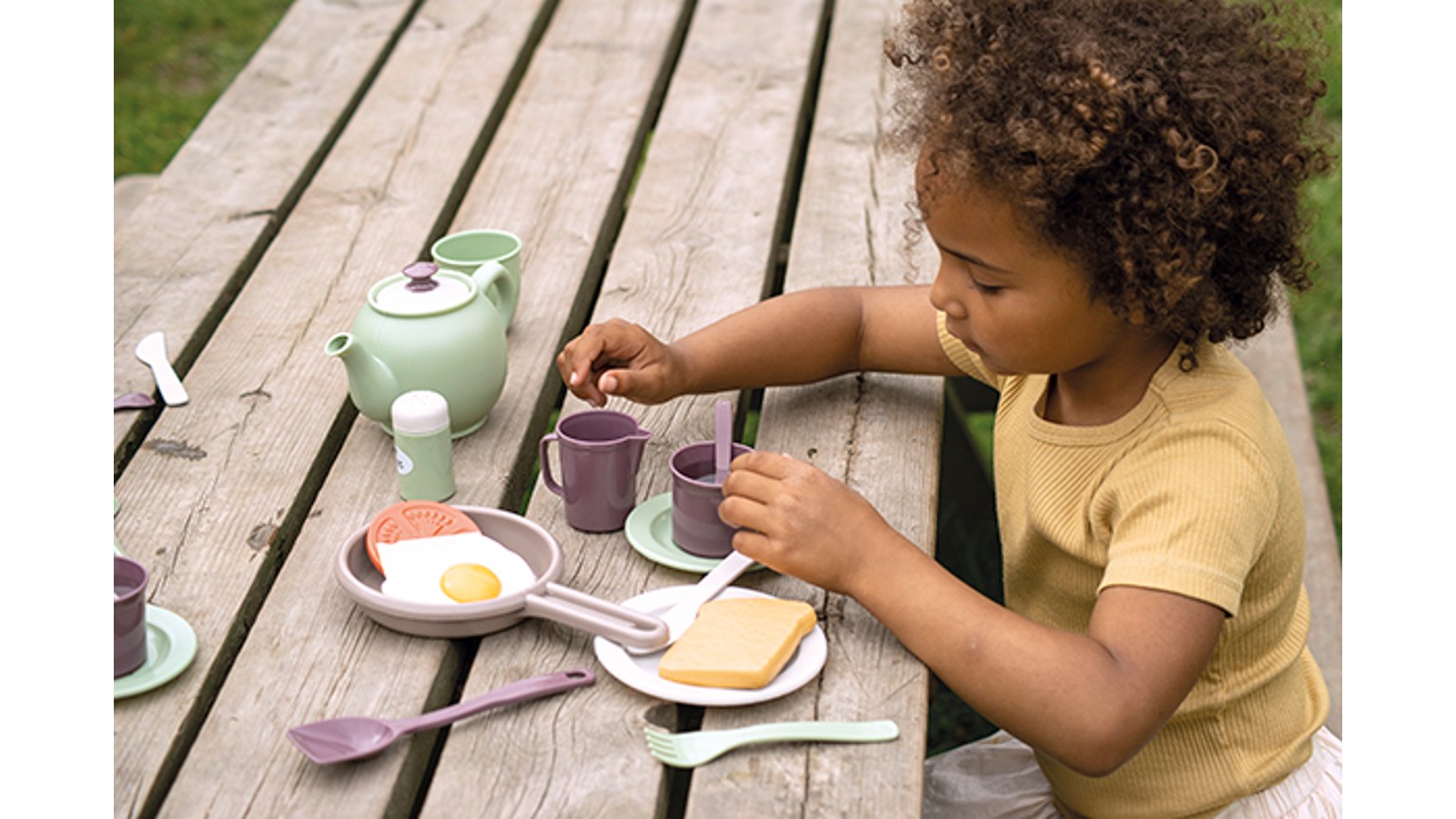 Barn ved udendørs bord med teselskab i lilla og mintgrønt, inklusiv kande og tallerkener.