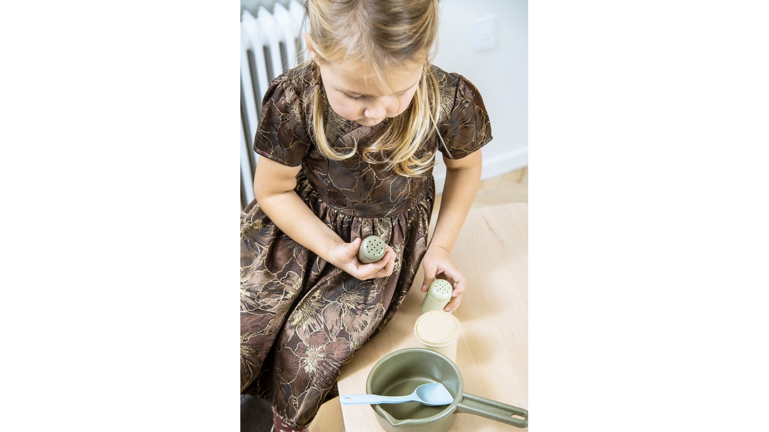 Barn i brun kjole leger med små køkkenredskaber over en skål på gulvet.
