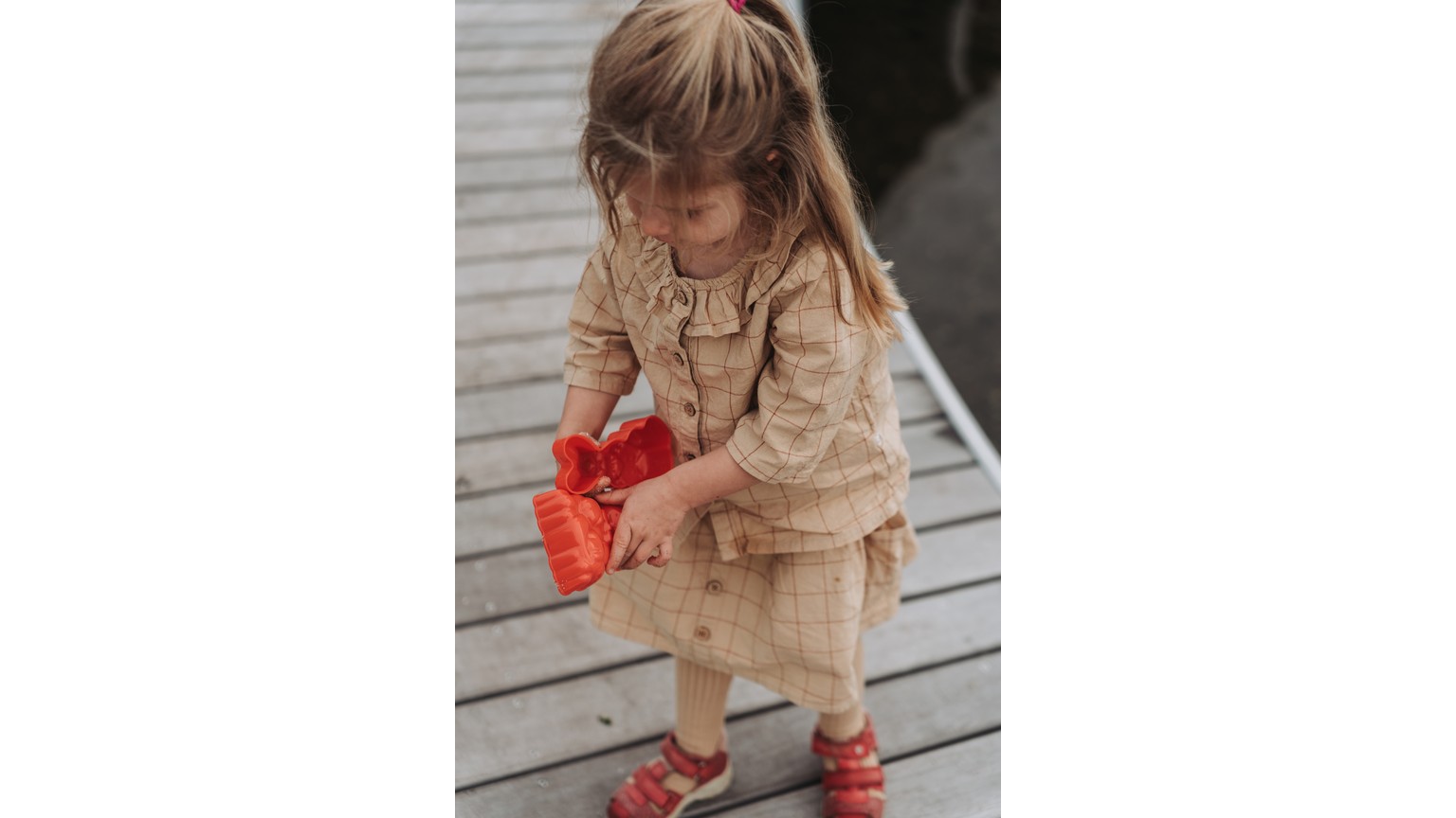 Barn i beige tøj og røde sko står på trædæk med rød legetøjsgenstand i hænderne