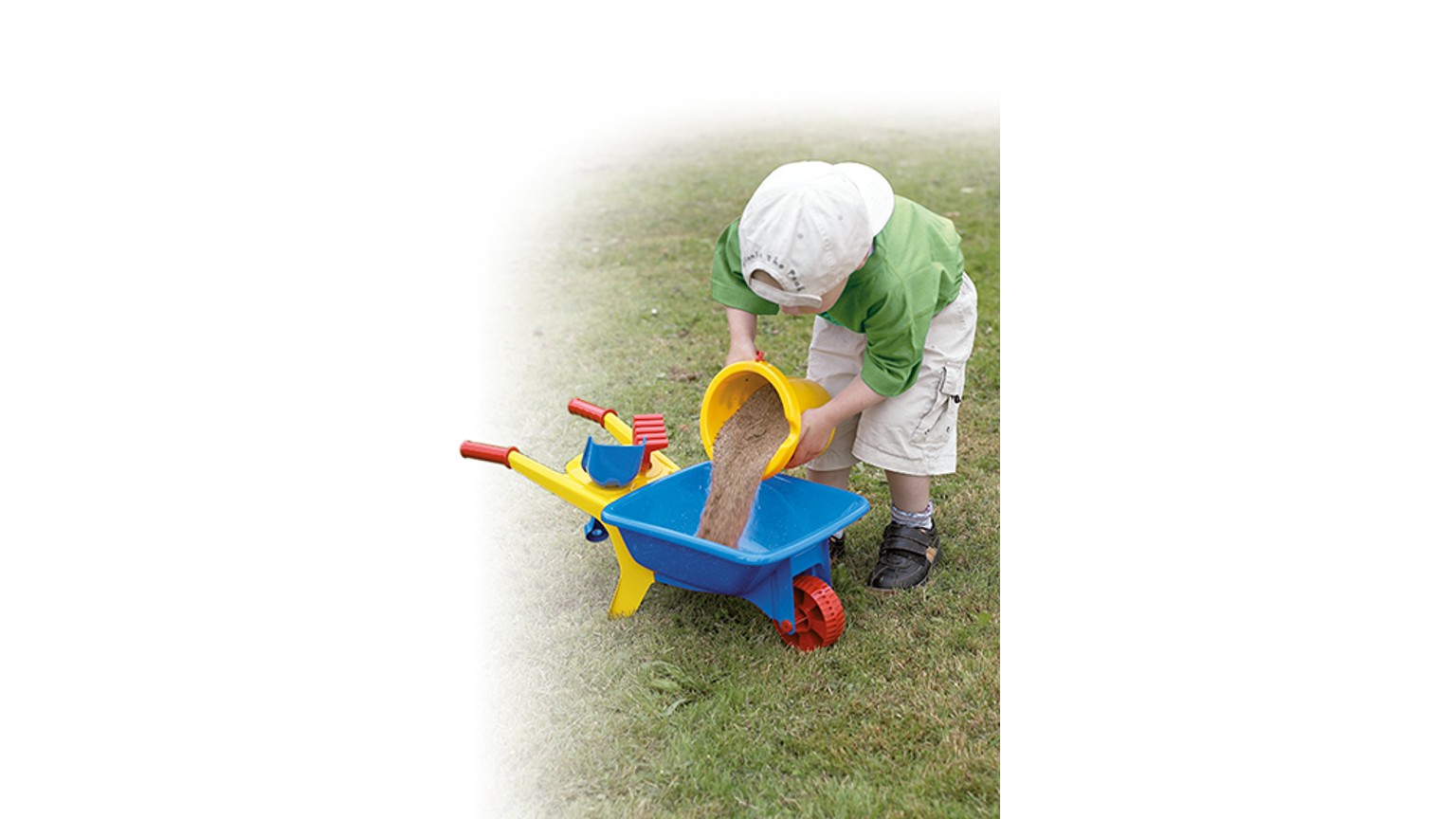 Et barn med grøn jakke hælder noget i en blå og gul trillebør på græsplæne.