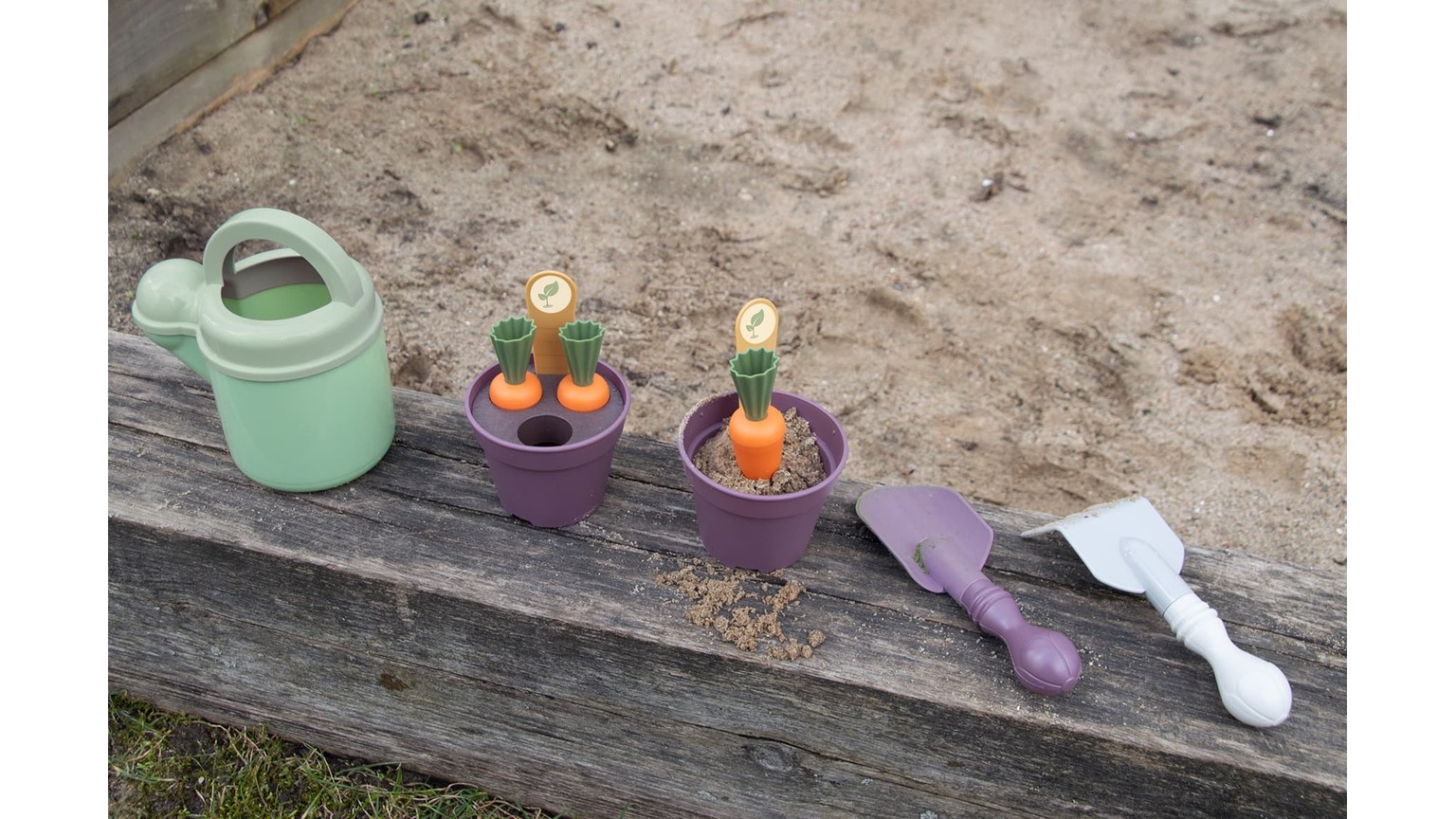 Grøn vandkande og to lilla plantepotter på træplanke med sand i baggrunden.