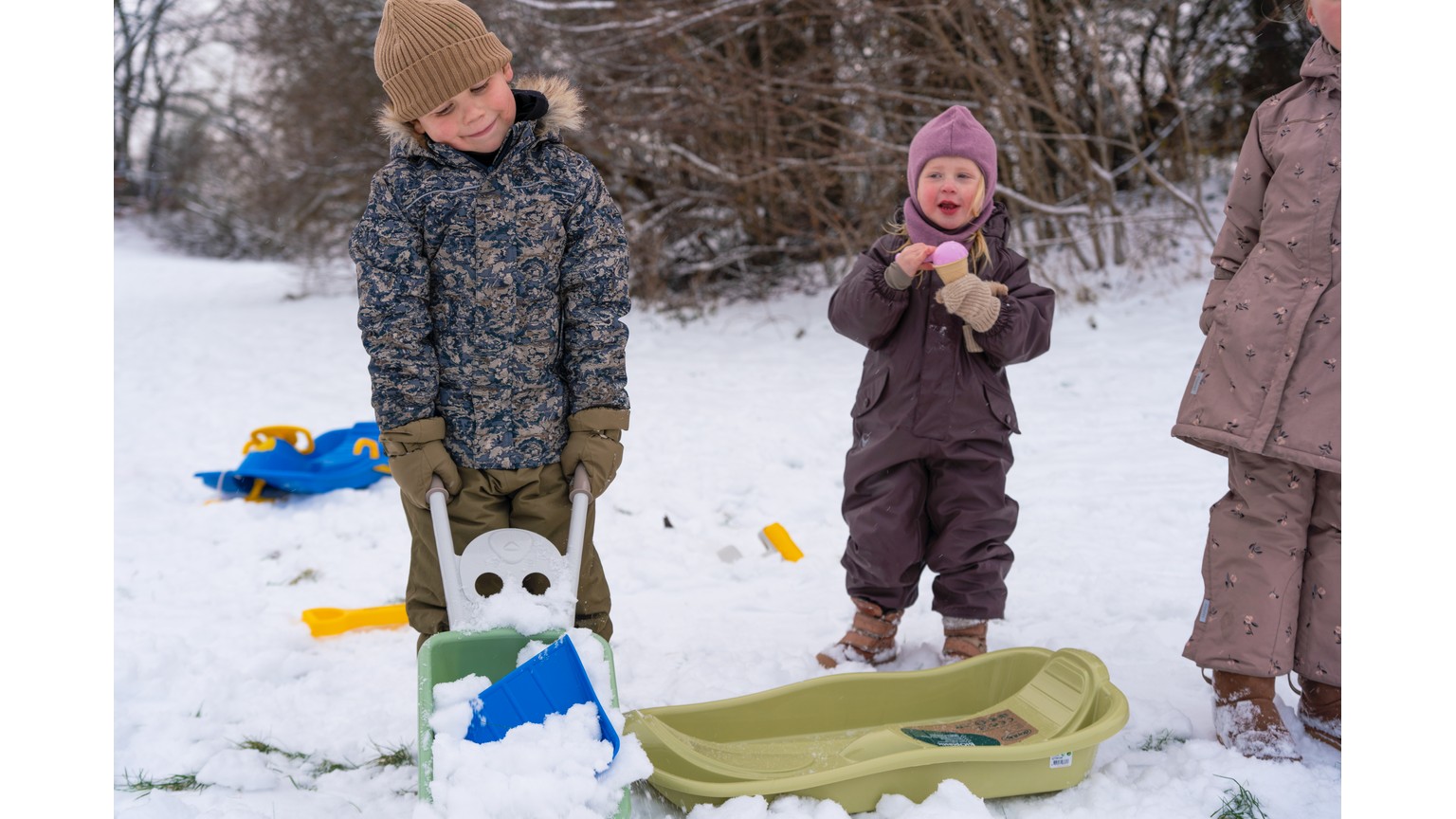 Børn med grønne og blå slæder i snelandskab på vinterdag.