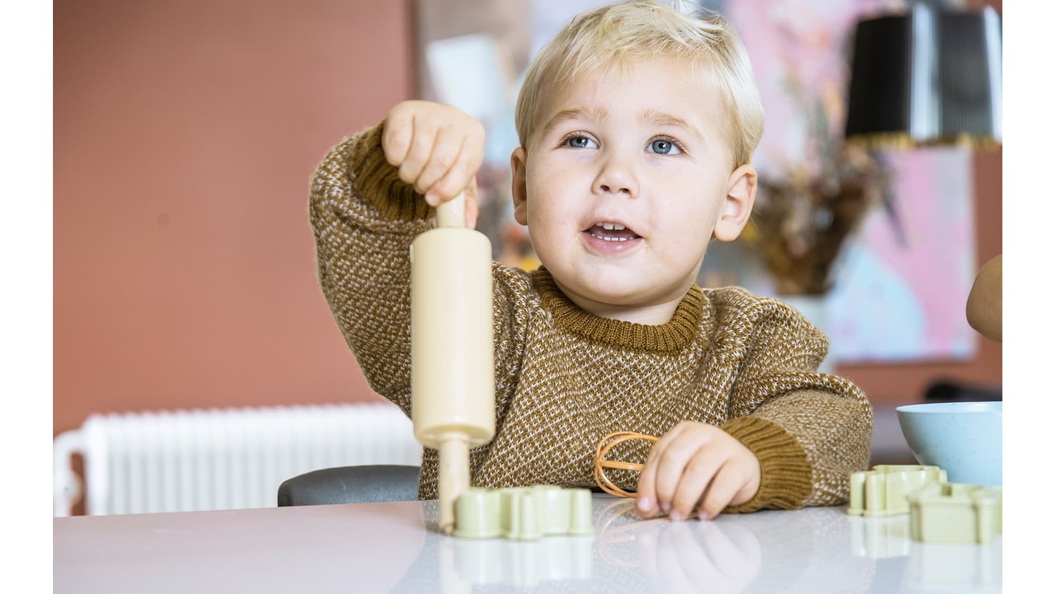 Lille barn i brun trøje sidder ved bord med bageredskaber og former i naturlige farver.