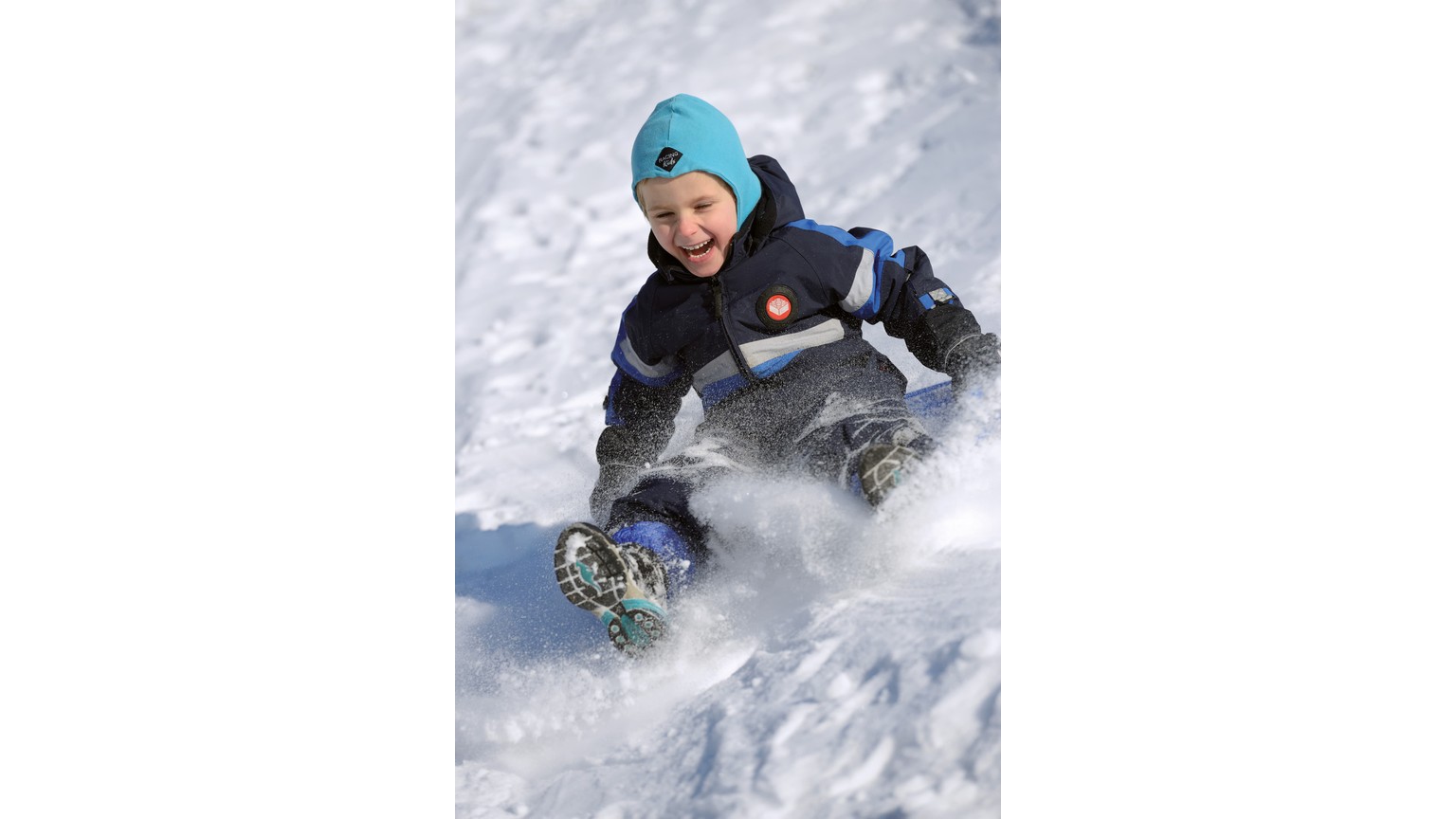 En person i vinterdress glider ned ad en snedækket bakke på en blå kælk i liggende position.