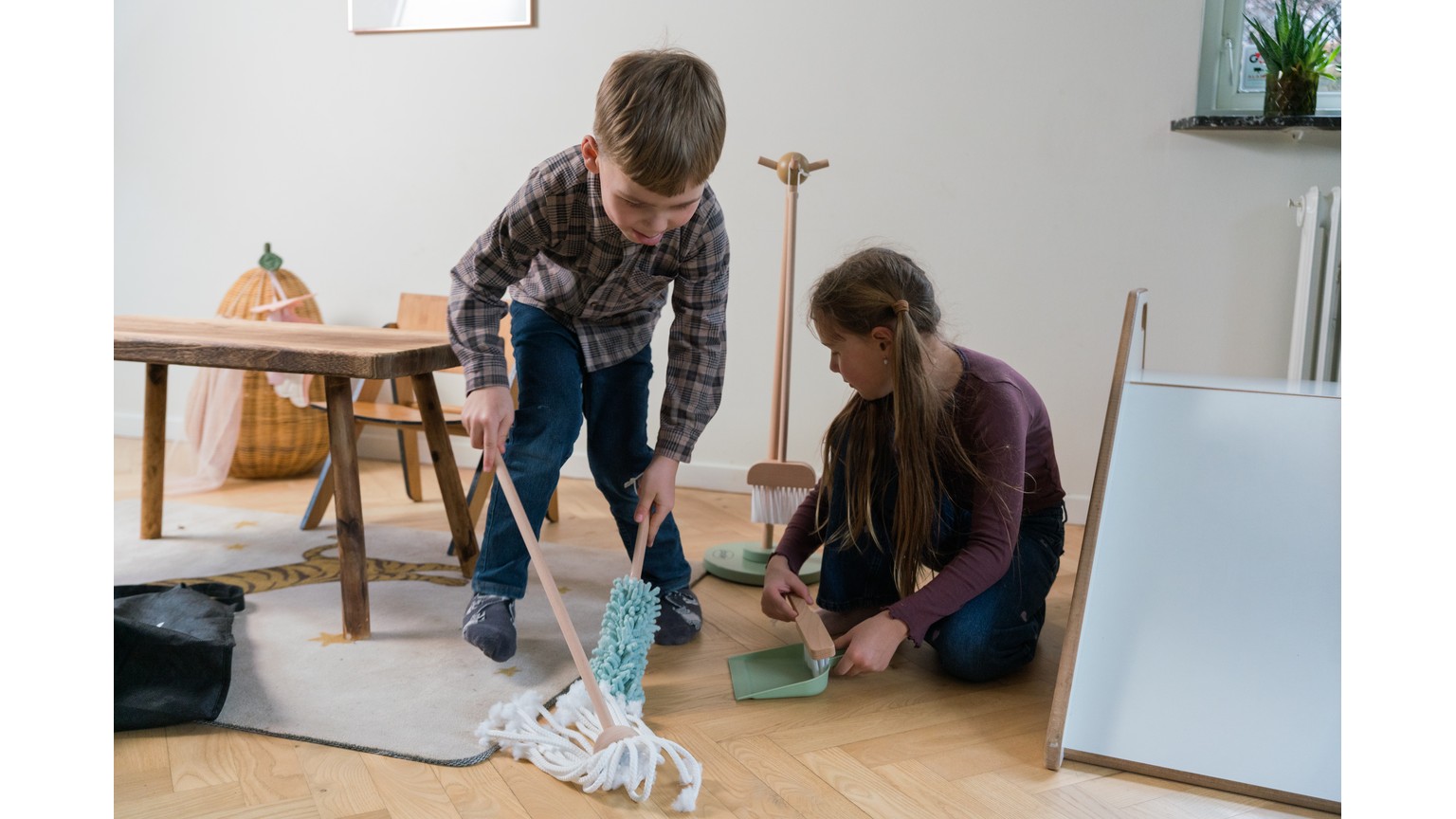 To personer gør rent på gulvet med moppe og fejebakke i hjemlig stue med vindueskarm og træbord.