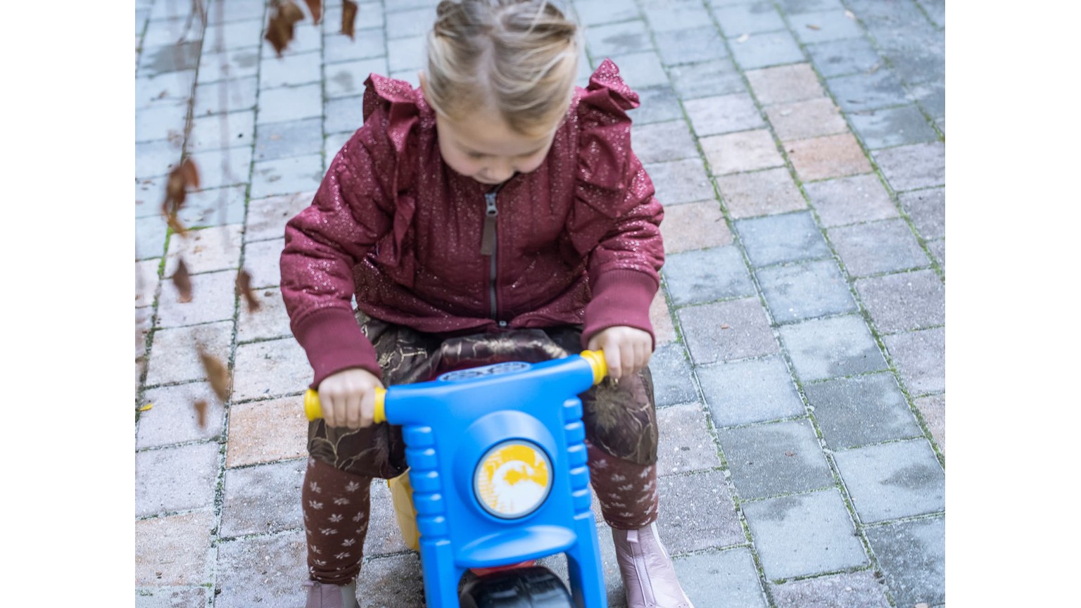Barn på blå scooter-legetøj på en flisebelagt gård