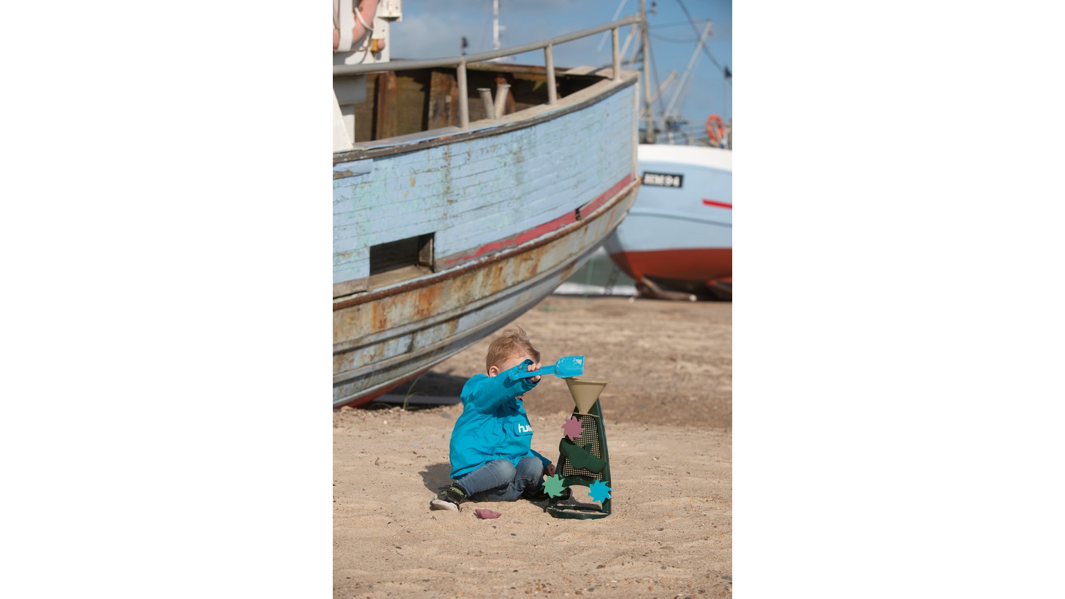 Person i turkis tøj sidder ved gamle fiskebåde og leger med blåt sandlegetøj på stranden.