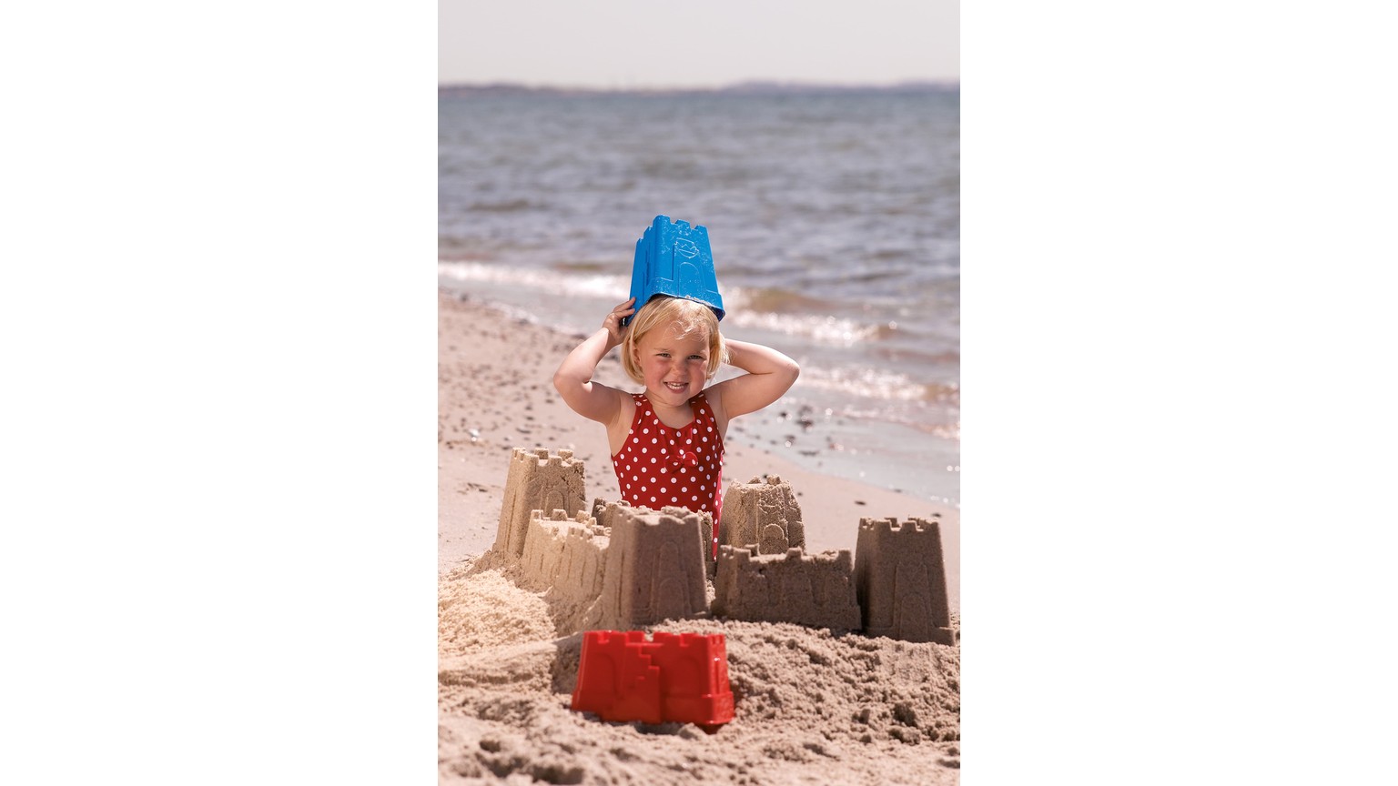 Et barn med blå hat bygger sandslot på stranden med rød spand og sandlegetøj ved vandkanten.