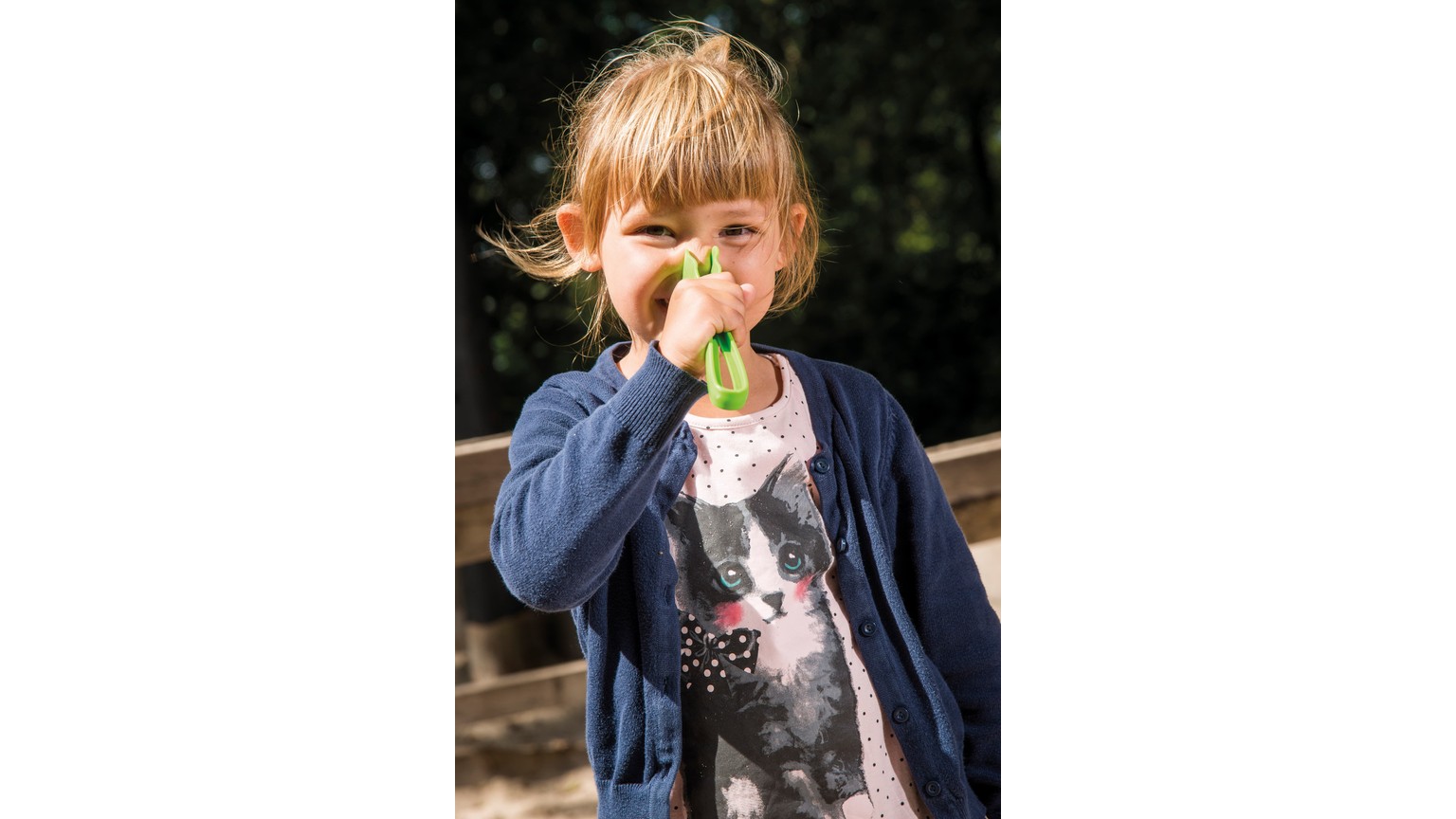 Barn med grøn legemadsdel ved næsen, iført mørkeblå cardigan i solskinsvejr