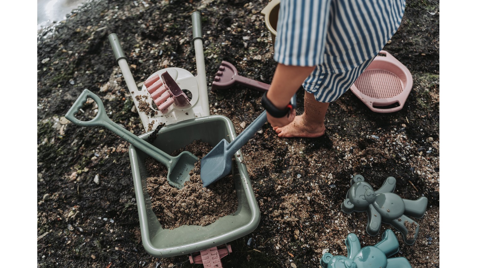 Person leger med grøn sandkasse-legetøjssæt på mørk jord med trillebør og haveredskaber