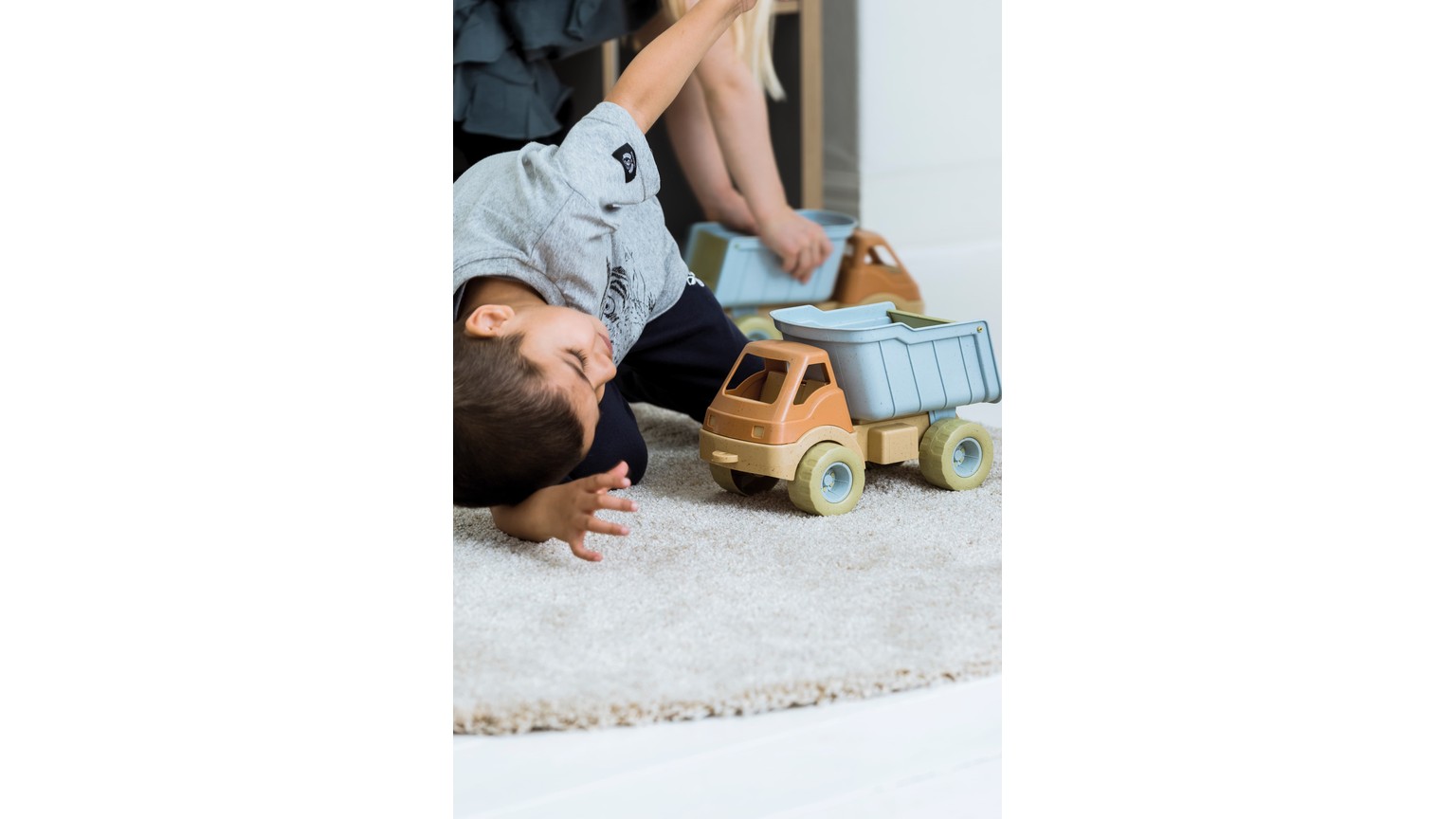 Person ligger på gulvtæppe ved siden af lyseblå og beige legetøjslastbil