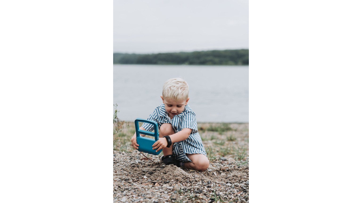 Lille barn med blåt sandlegetøj ved vandkant på en sommerdag.