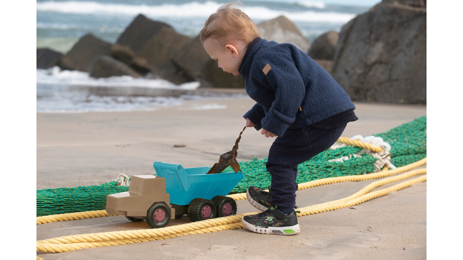 Barn leger med blå dumper på stranden ved siden af grønt legetøj og klipper.