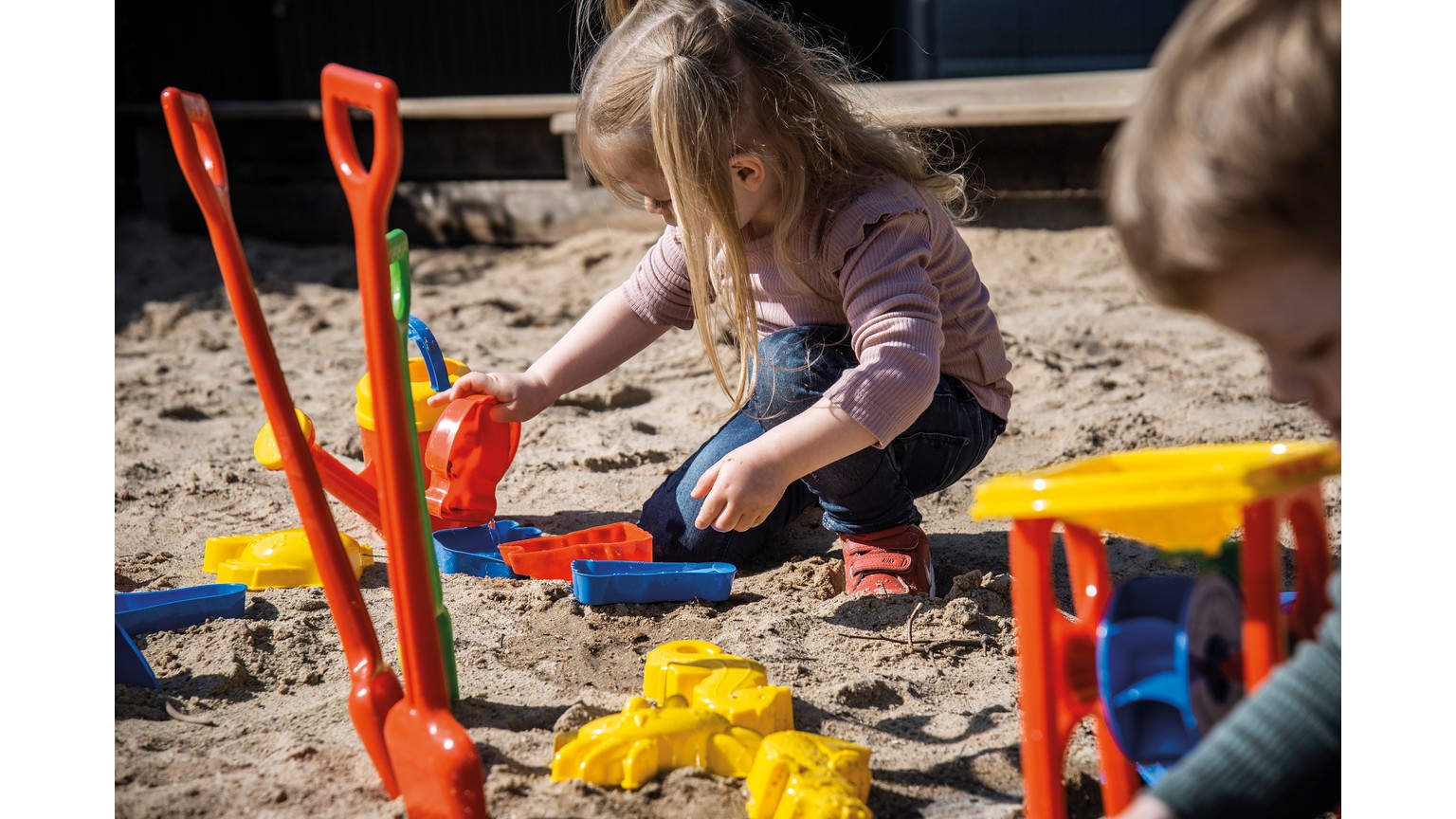 Barn leger med farverige sandlegetøj på sandområde med røde, gule og blå skovle og former.