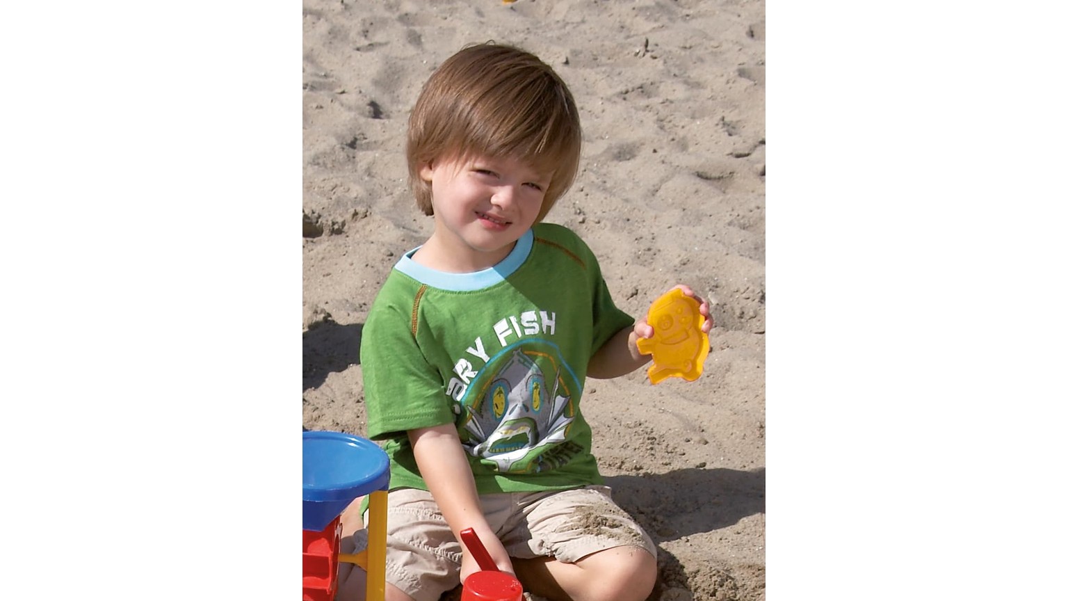 Barn i grøn t-shirt leger med gult sandlegetøj på stranden.