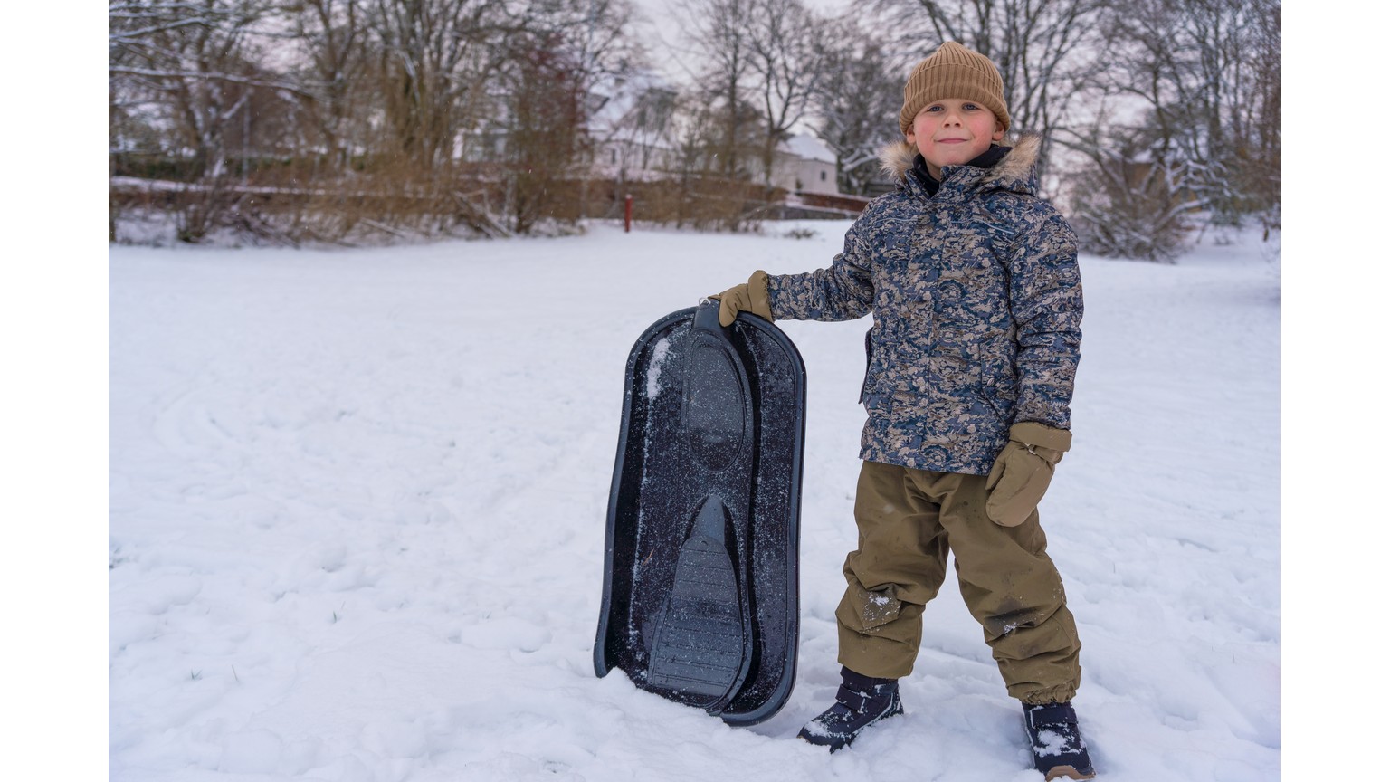 Person med kælk står i snedækket landskab på en vinterdag med træer i baggrunden.