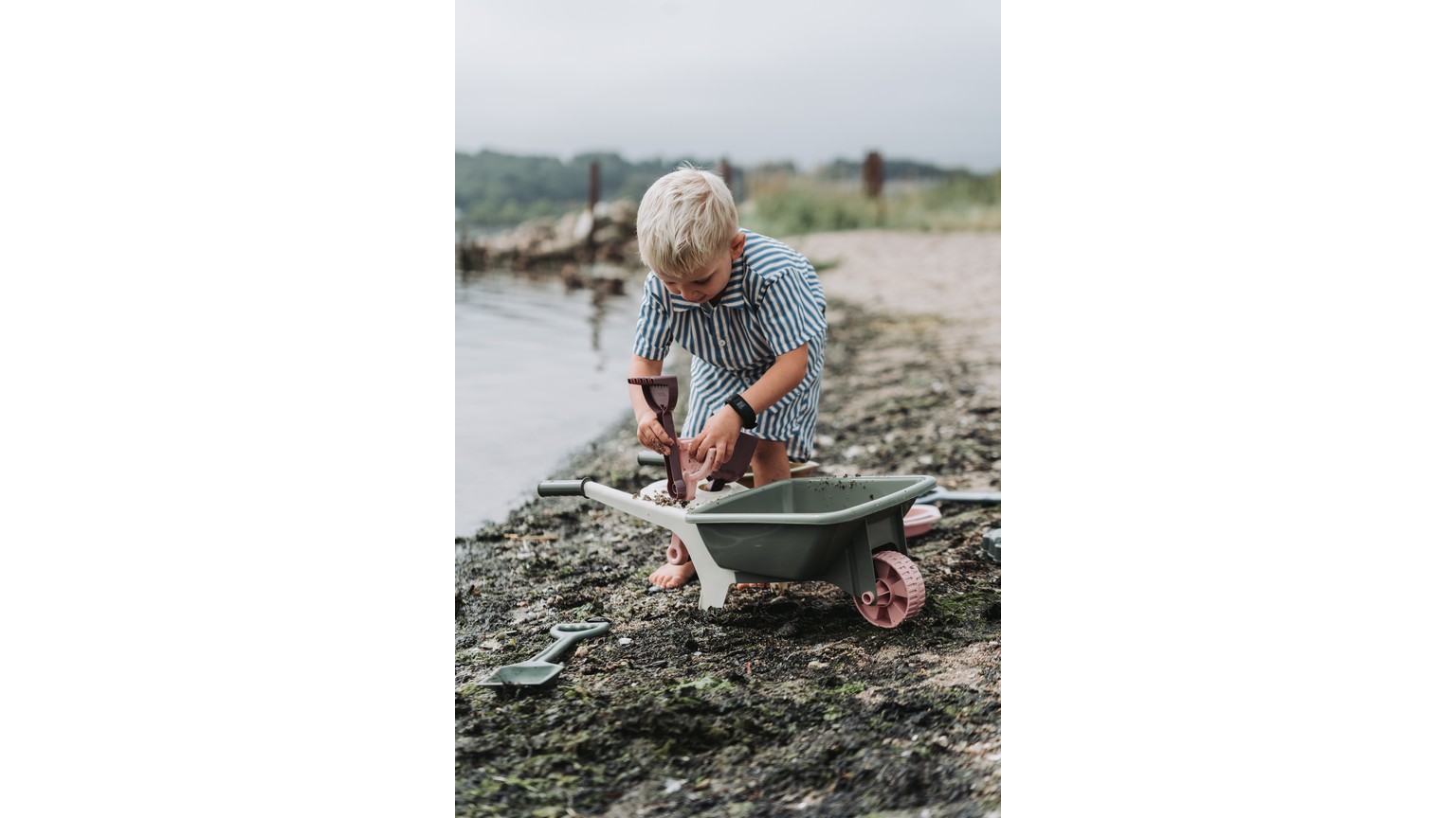 Barn med grøn trillebør leger ved stranden i gråvejr