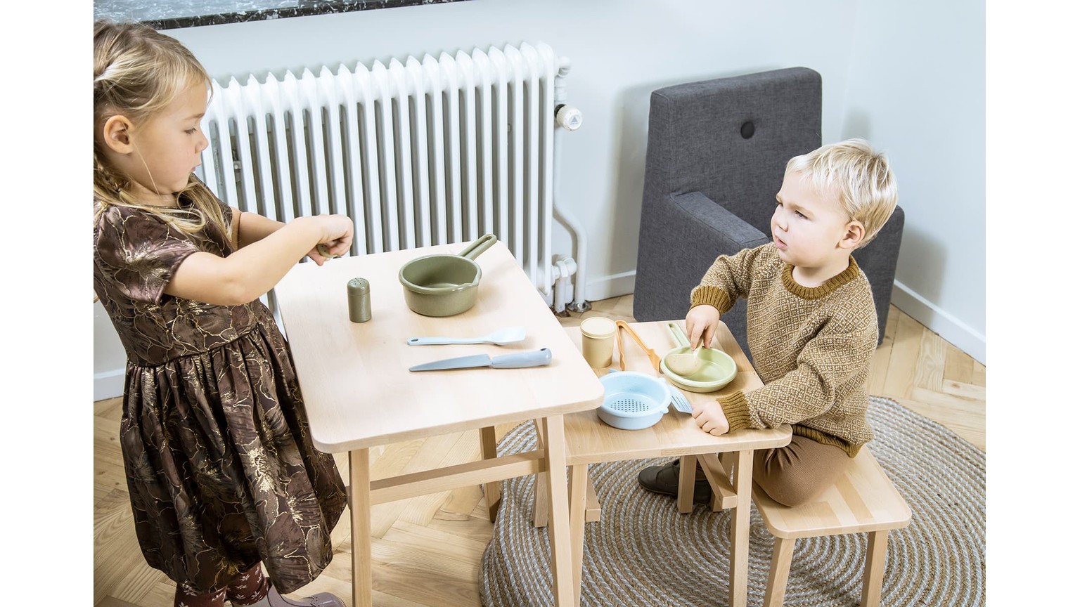 To børn sidder ved et lille bord med legemad og tallerkener i et rum med en radiator på væggen.